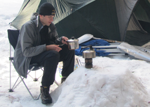 Alex Mitchell's snow table: a camper piled up snow to make a small table and has his stove on it, pan in hand to cook