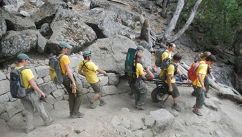 nps photo by Jeremy Bernfeld litter on Yosemite Falls trail: six people hold the sides of a wheeled litter bringing a victim down a trail, two other Yosemite Search and rescue people behind the litter