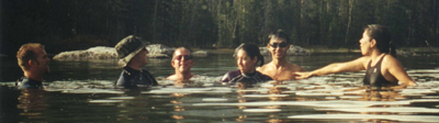 swimminginStringLake400pixel: a group of people in swimsuits and wetsuit standing in a mountain lake, reacting to a joke