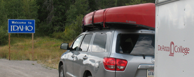 welcome to Idaho and college gear hauler trailer: Along a highway, a welcome to Idaho sign, and the De Anza College gear hauler trailer towed by a car with a canoe on top