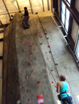 Alex and Alanna at climbing gym: girls belays guy nearing top of climbing wall