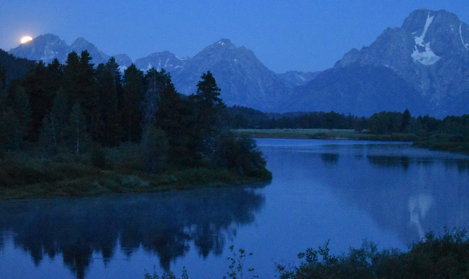 mountains with part of full moon behind them, water in the foreground