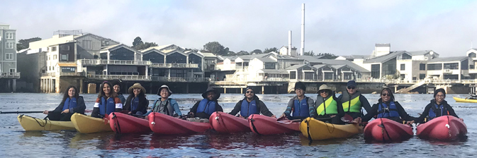 row of kayakers with Monterey Bay Aquarium behind them