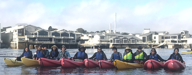 row of kayakers with Monterey Bay Aquarium behind them