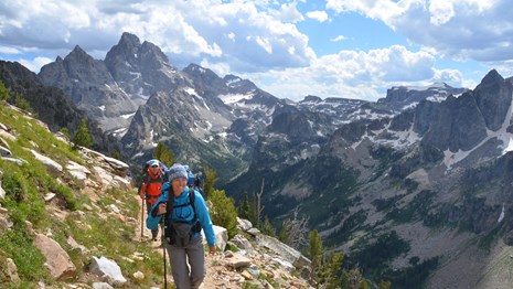 2 people with backpacks on a trail with peaks behind them