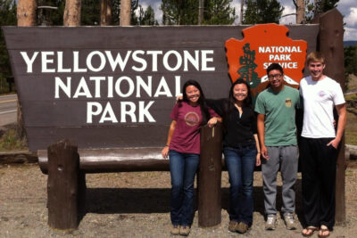 4 people standing in frontt of a large wooden sign that says Yellowstone National park