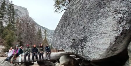 seven people sitting on a fallen tree next to a huge glacial erratic