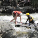 man wearing a helmet and lifejacket crawling across a ladder laid out for him by rescuers across a rocky stream full of whitewater