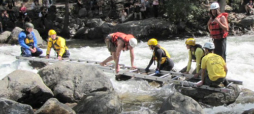 man wearing a helmet and lifejacket crawling across a ladder laid out for him by rescuers across a rocky stream full of whitewater